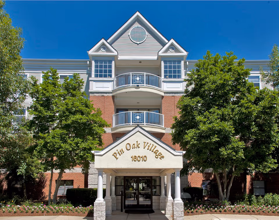 Front entrance of the Pin Oak Village building with a covered portico, balconies above, and trees framing the facade.