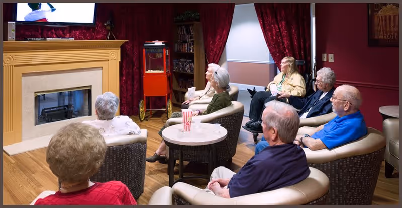 Elderly residents sit in a cozy senior living lounge watching TV near a fireplace and popcorn machine.