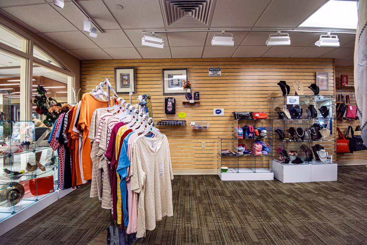 Interior view of a retail area in a senior living facility with clothing racks displaying various shirts and blouses, glass display cases with jewelry and accessories, and handbags hanging on the wall. The space has carpeted flooring, a wooden slat wall, and ceiling track lighting.