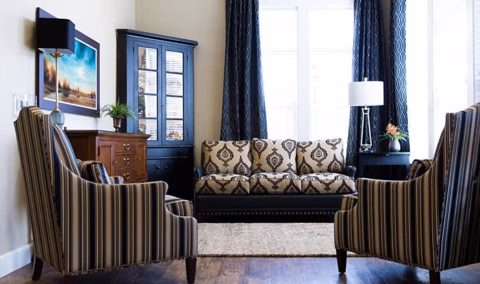 A well-lit living room with a patterned sofa centered against a window with dark blue curtains. Two striped armchairs face the sofa. There is a wooden cabinet with glass doors and a chest of drawers with a lamp and a small plant on top. A framed landscape painting hangs on the wall.