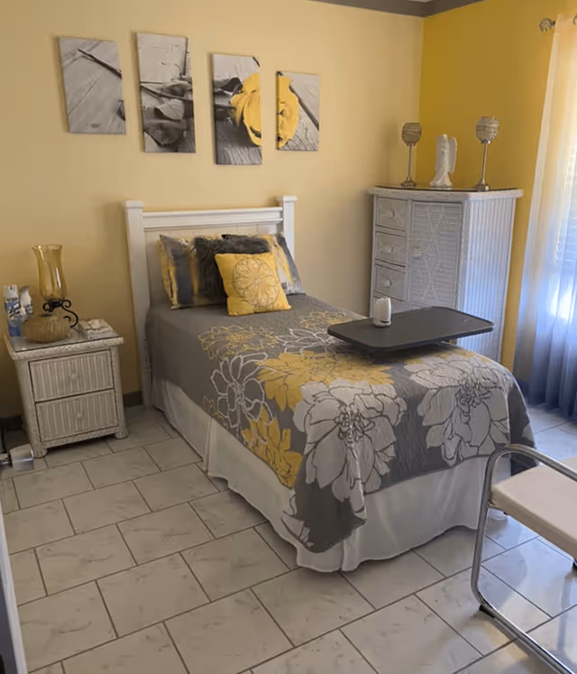 A neatly made single bed with a gray and yellow floral bedspread and matching pillows in a small bedroom. The room has light yellow walls, a white wicker nightstand with a yellow glass lamp and other small items, and a white wicker dresser with decorative items on top. A window with sheer curtains allows natural light into the room. The floor is tiled with light-colored tiles.