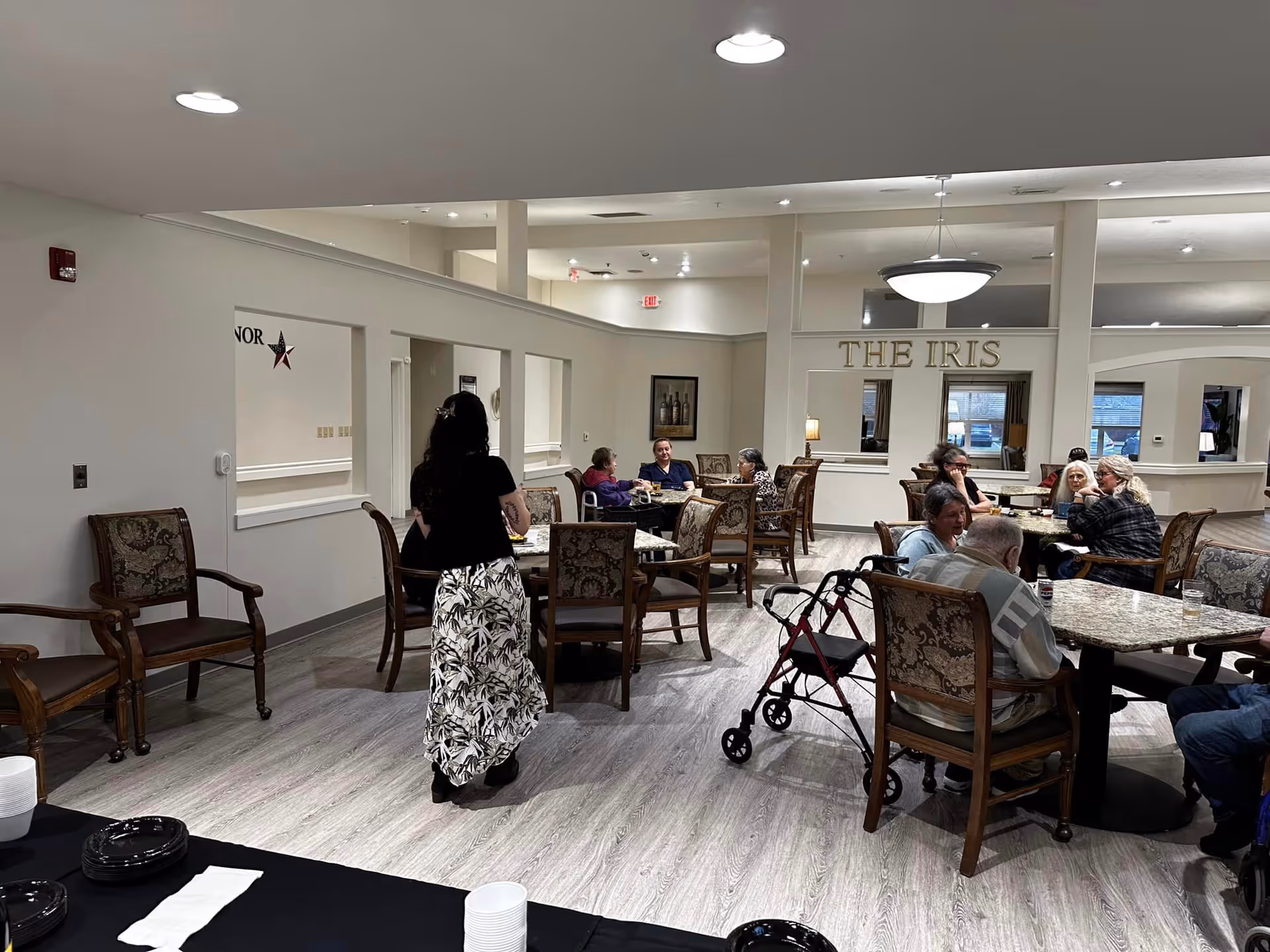 A communal dining area in The Iris Memory Care facility with several elderly residents seated at tables, some engaged in conversation. A woman in a black top and patterned skirt is walking through the room. The room has wooden chairs with patterned upholstery, marble-top tables, and light-colored wood flooring. The wall in the background has the words 'THE IRIS' displayed above windows looking into another room.