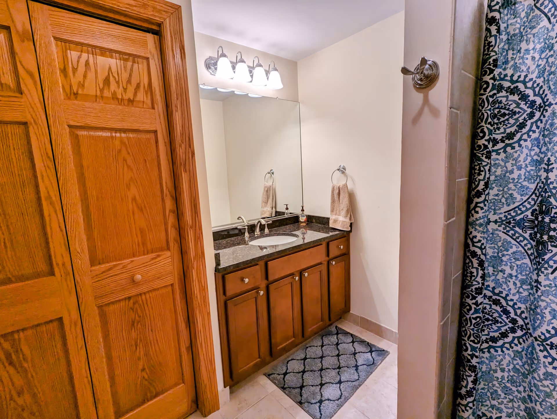 Bathroom with wooden cabinets and a granite countertop sink. Above the sink is a large mirror with four light fixtures. There is a towel hanging on a ring on the wall and a patterned blue and white shower curtain visible on the right side. A wooden closet door is on the left side of the image.