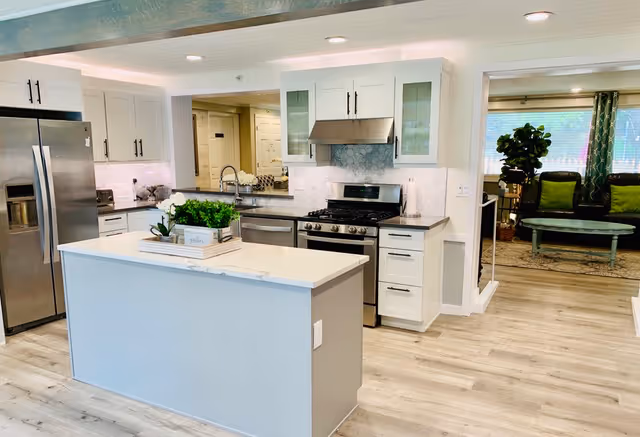 Modern kitchen with a large island in the center, white cabinetry, stainless steel refrigerator, stove with hood, and a sink. The kitchen opens into a living area with a green couch and a large window with curtains.