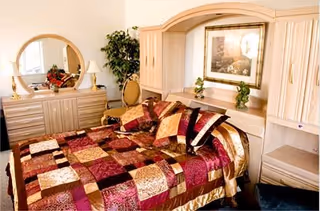 A cozy bedroom featuring a bed with a colorful patchwork quilt in shades of red, gold, and brown. The room includes light wood furniture such as a dresser with a round mirror, a built-in headboard with cabinets, a framed picture above the bed, and a small plant on the dresser. A chair and a potted plant are also visible in the corner.