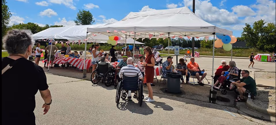 Outdoor gathering at Forest View Manor with seniors and caregivers under white canopy tents decorated with balloons and small American flags. People are sitting and standing around tables covered with red and white striped tablecloths, enjoying a sunny day in a parking lot area.