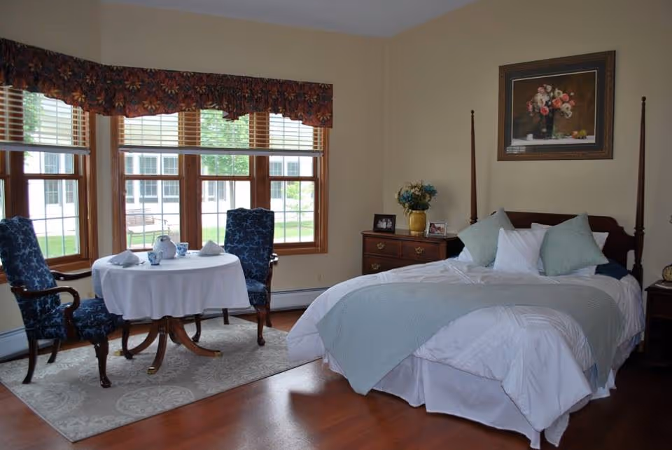 A cozy bedroom with a large bed covered in white and light blue bedding, two blue patterned chairs around a small round table with a white tablecloth, and large windows with wooden frames and floral valances letting in natural light. There is a wooden dresser with a vase of flowers and framed photos, and a floral painting hanging on the wall above the bed.