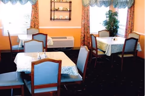 A dining room with several tables covered with patterned tablecloths, each surrounded by wooden chairs with blue cushions. The walls are painted peach with white trim, and there are windows with floral curtains and blue valances. A small shelf with decorative plates is mounted on the wall, and a potted plant is placed near one of the windows.