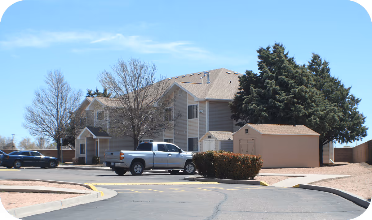 Two-story senior living facility exterior with parked trucks, trees, and a beige shed