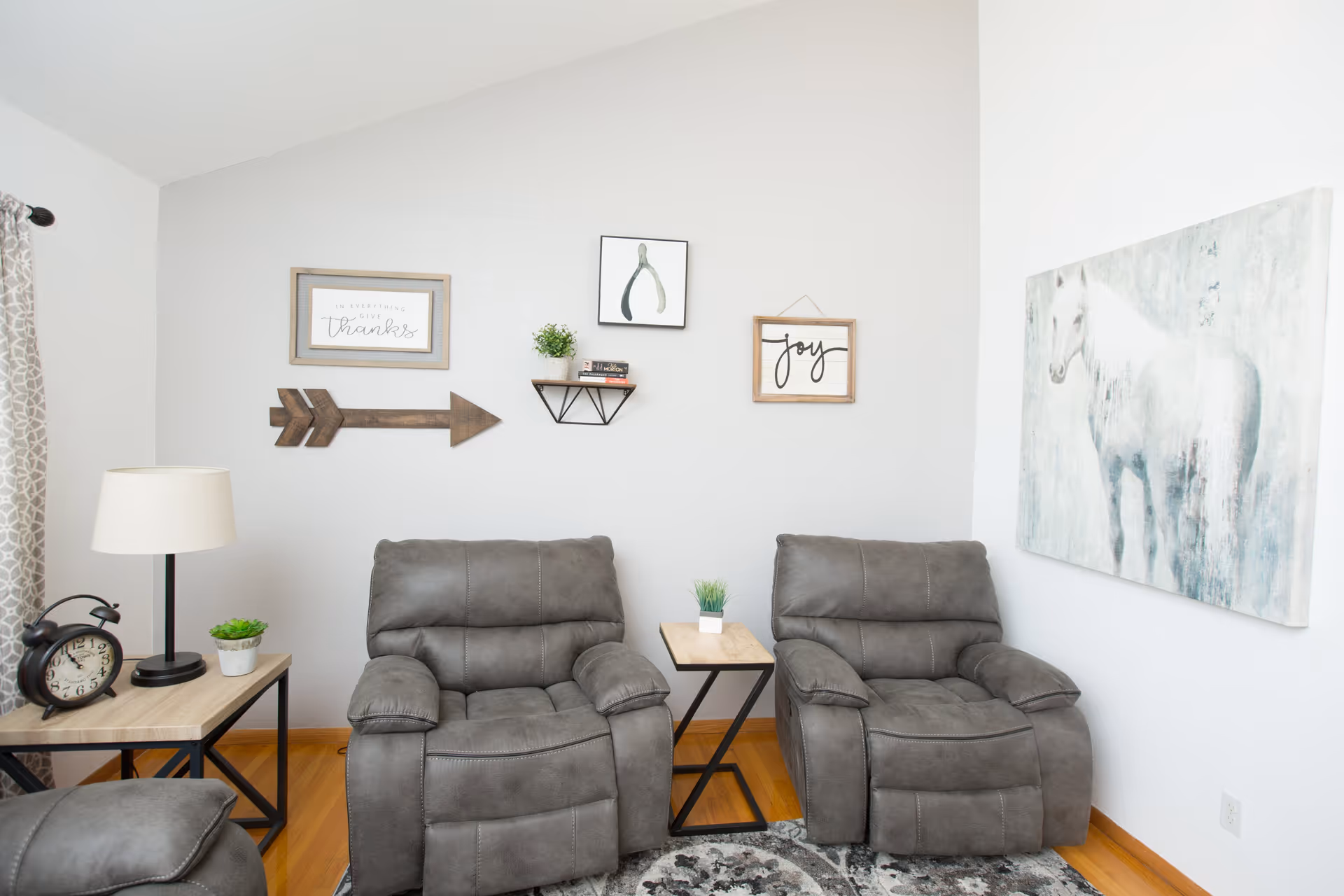 A cozy living room area with two gray recliner chairs separated by a small wooden side table with a small plant. On the left, there is a wooden side table with a lamp, a small plant, and a vintage-style alarm clock. The walls are decorated with framed art including a white horse painting, a wooden arrow, and signs that say 'joy' and 'in everything give thanks'. The floor has a patterned rug and the room has light-colored walls and wooden flooring.