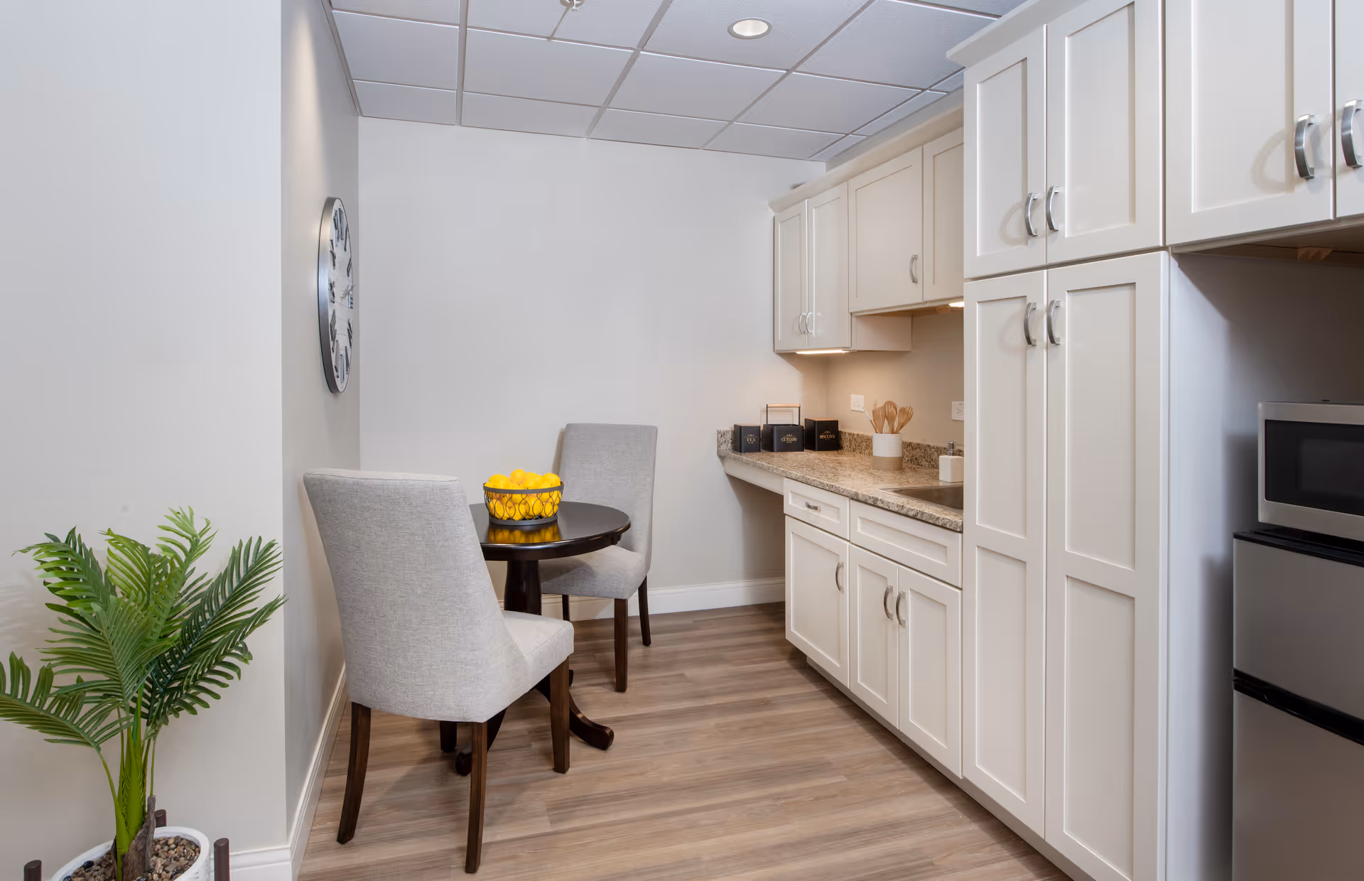 Small kitchenette with white cabinets, a granite countertop, and a round table with two upholstered chairs and a bowl of lemons.