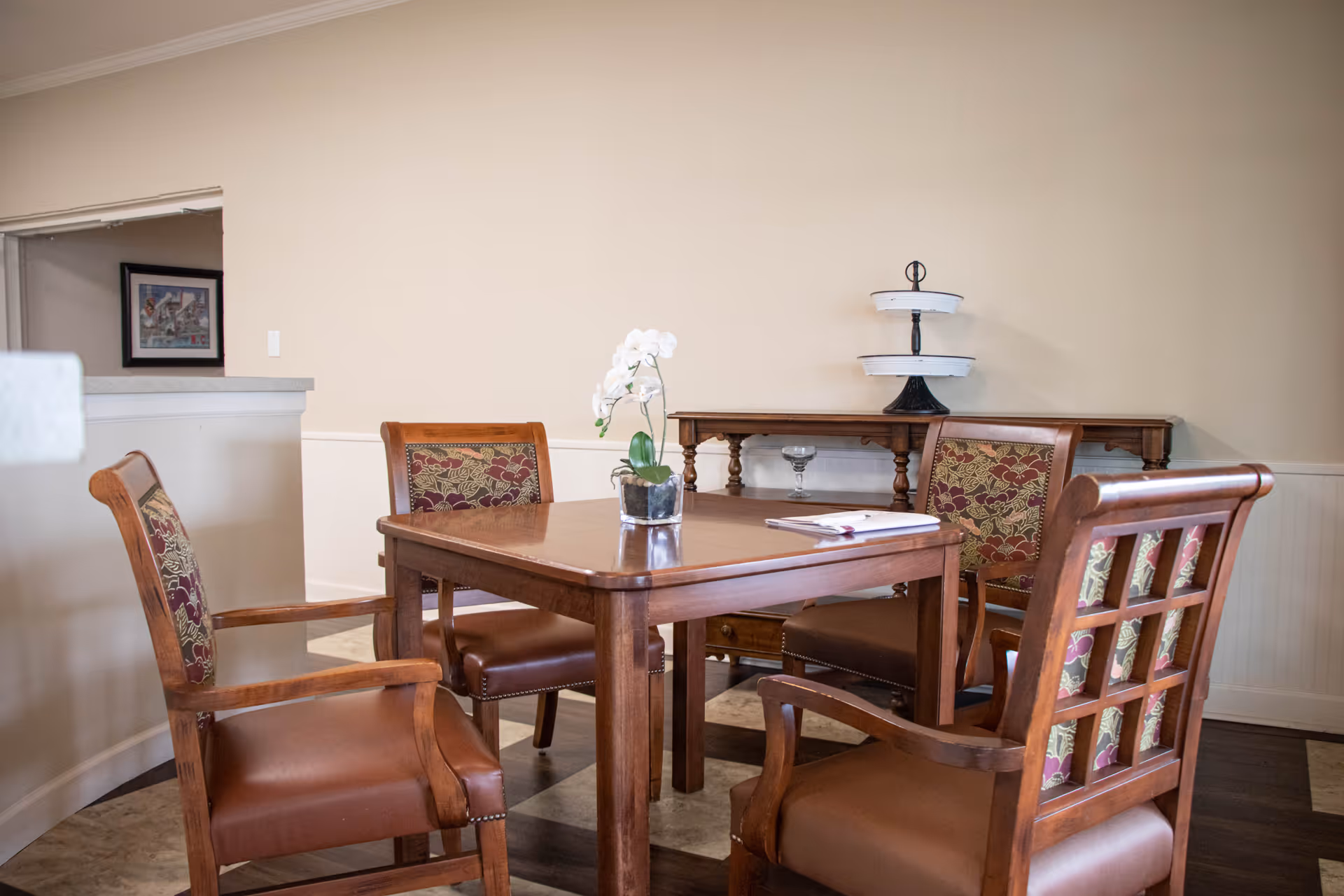 A small dining area with a wooden square table surrounded by four wooden chairs with patterned upholstery. On the table is a small potted white orchid and a closed notebook with a pen. In the background, there is a wooden sideboard with a two-tiered serving tray and a glass bowl. The walls are beige and the floor has a patterned design.