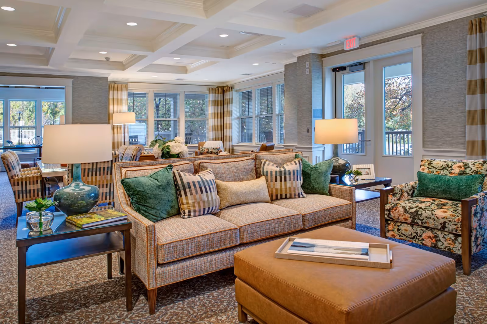 Bright, comfortable common room with upholstered sofas, armchairs, an ottoman, side tables, lamps, and large windows.