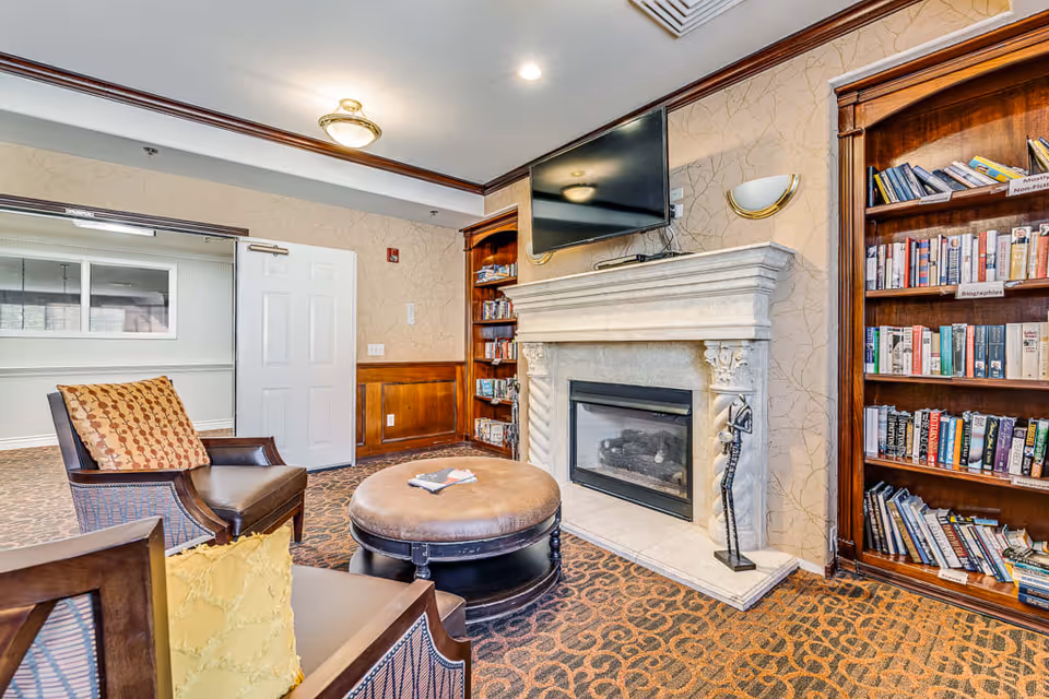 A cozy senior living room featuring two cushioned armchairs with patterned pillows, a round ottoman in the center, a stone fireplace with a mounted flat-screen TV above it, and wooden bookshelves filled with books on either side of the fireplace. The room has patterned carpet flooring, beige wallpaper, and a white door leading to another room.