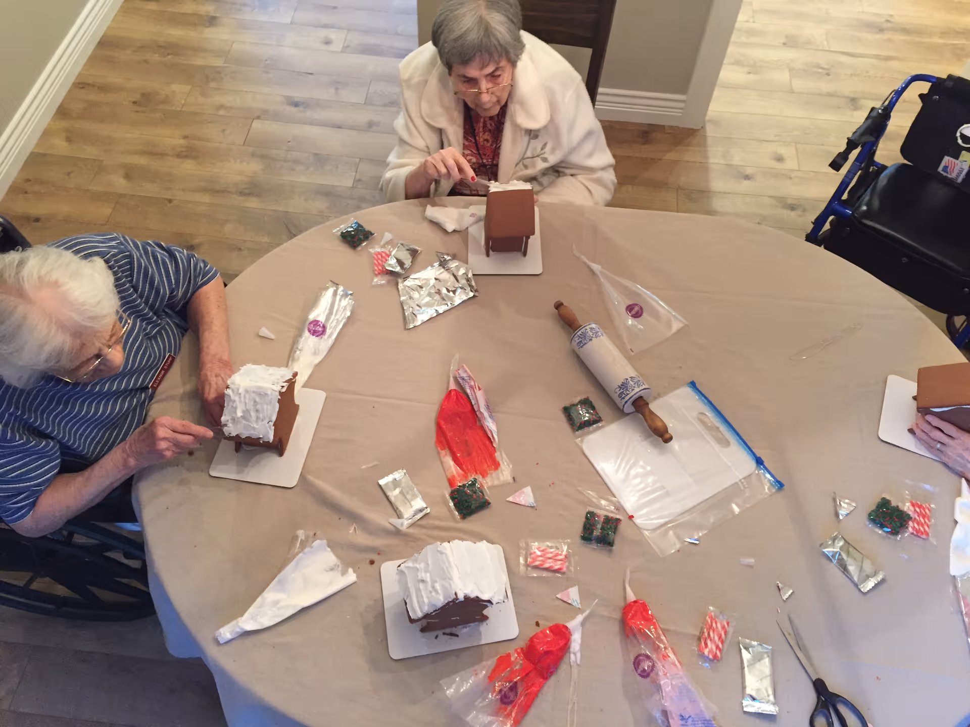 Three elderly women sitting around a round table covered with a beige tablecloth, decorating small gingerbread houses with white icing and various colorful candies. There are decorating tools such as piping bags, a rolling pin, and scissors on the table. One woman is in a wheelchair, and the floor is wooden.