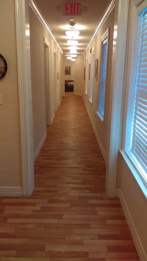 A long, narrow hallway with wooden flooring and cream-colored walls. There are several doorways on the left side and windows with blinds on the right side. Ceiling lights are evenly spaced along the hallway, and an illuminated red exit sign is visible at the far end. A couch and framed pictures can be seen at the end of the hallway.