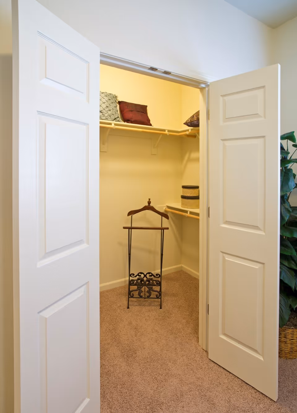 Open closet with beige carpet and cream-colored walls. Inside the closet, there are two shelves with decorative pillows and storage baskets. A wooden valet stand with a metal base is positioned on the floor. The closet doors are white and partially open, and a green leafy plant in a woven basket is visible to the right outside the closet.