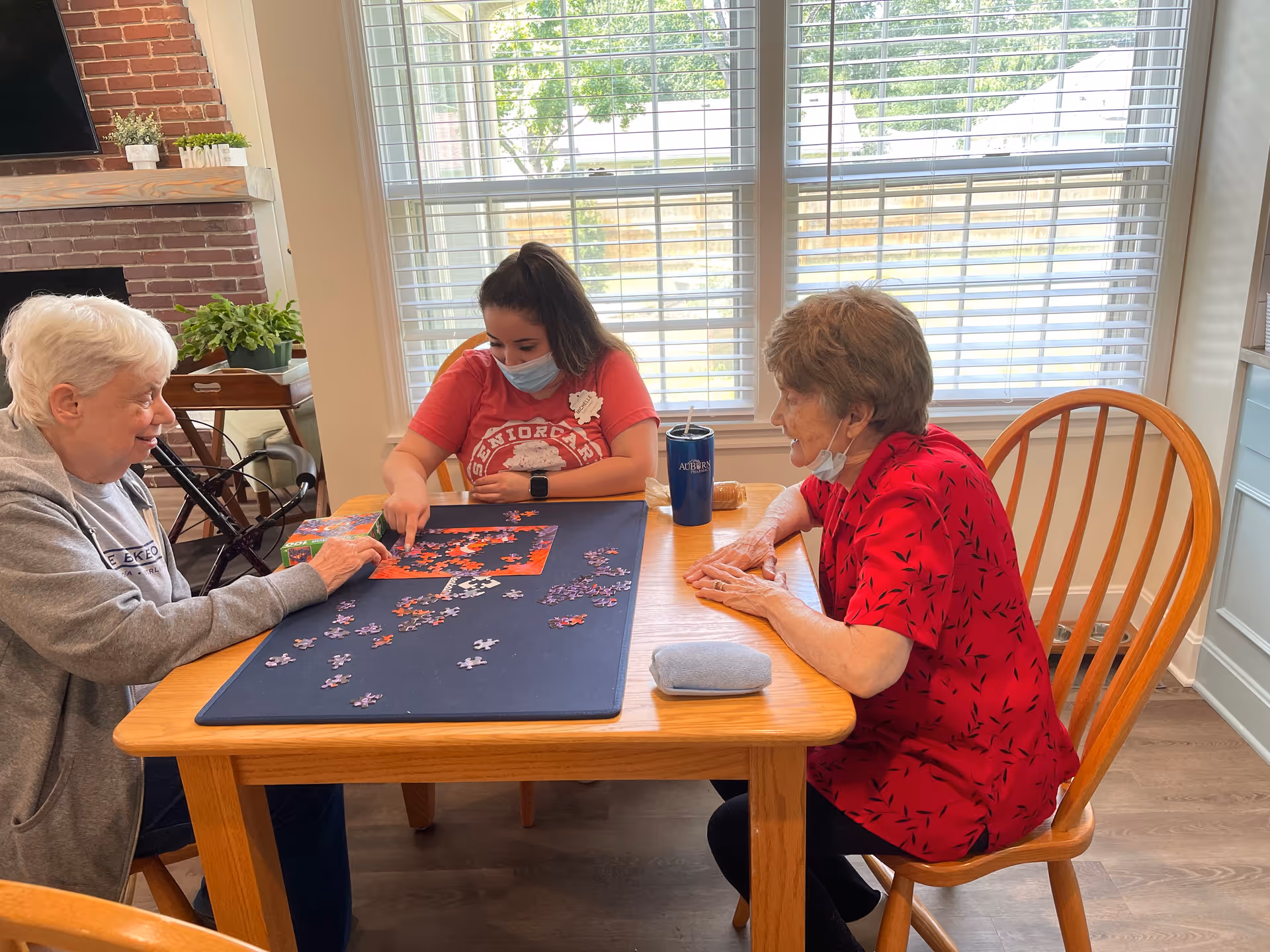 Three women sitting around a wooden table working on a jigsaw puzzle in a bright room with large windows. One woman is wearing a red shirt and a face mask, another woman in a gray sweatshirt, and the third woman in a red patterned blouse. There is a brick fireplace and a TV in the background.