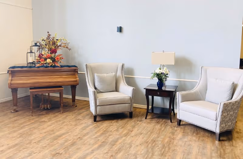A cozy seating area with two beige armchairs separated by a small wooden side table holding a lamp and a vase with flowers. To the left, there is a wooden piano adorned with a floral arrangement and decorative lanterns. The floor is wooden, and the walls are light-colored.