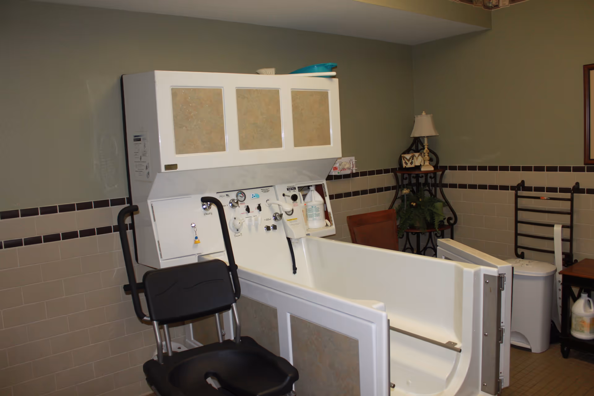 A senior bathing room featuring a walk-in bathtub with control knobs and a black shower chair. The room has beige tiled walls with a dark brown border, a small table with a lamp and decorative items, and a plant stand with greenery.