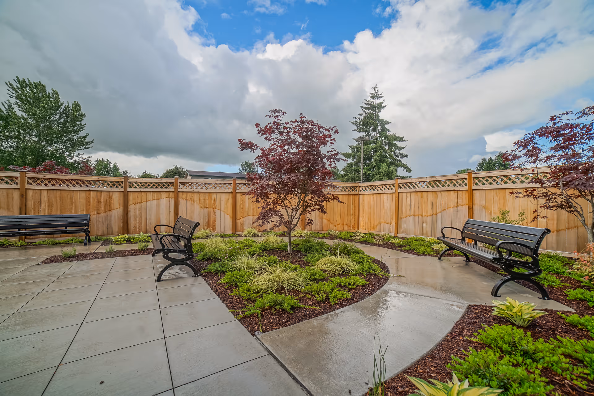 Walled courtyard with benches, landscaped garden beds, a small tree and wet paved pathways under a cloudy sky.