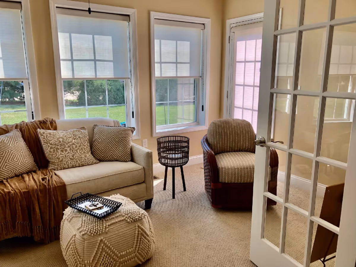 A cozy living room with a beige sofa adorned with patterned cushions and a brown throw blanket. There is a textured beige ottoman with a black tray on top in front of the sofa. Next to the sofa is a wicker armchair with striped upholstery. The room has three large windows with white blinds partially drawn, letting in natural light. A glass-paneled door is open, revealing part of the room.