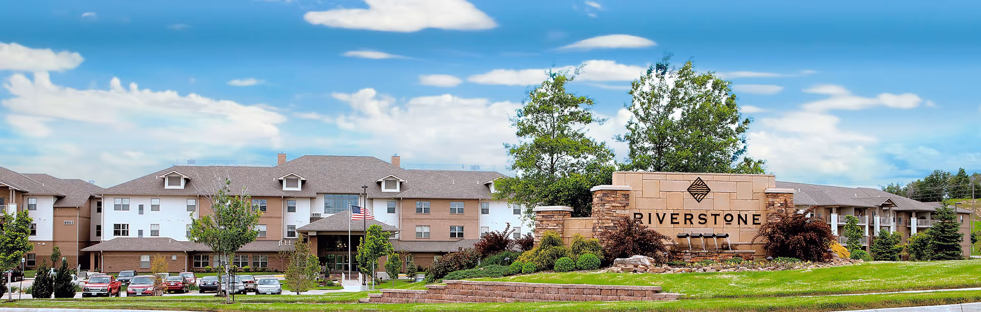 Wide exterior view of Riverstone Retirement Community showing a large multi-story building with a brown and white facade, surrounded by green lawns, trees, and a parking lot with several cars. A prominent stone sign with the name 'Riverstone' is in the foreground, under a partly cloudy blue sky.