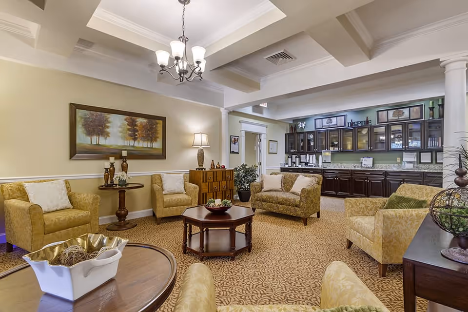 A cozy living room area in a senior living facility featuring multiple upholstered armchairs and sofas arranged around wooden coffee tables. The room has warm beige walls, a patterned carpet, a chandelier hanging from a tray ceiling, and a large framed painting of trees on the wall. In the background, there is a kitchenette area with dark wood cabinets and a granite countertop.