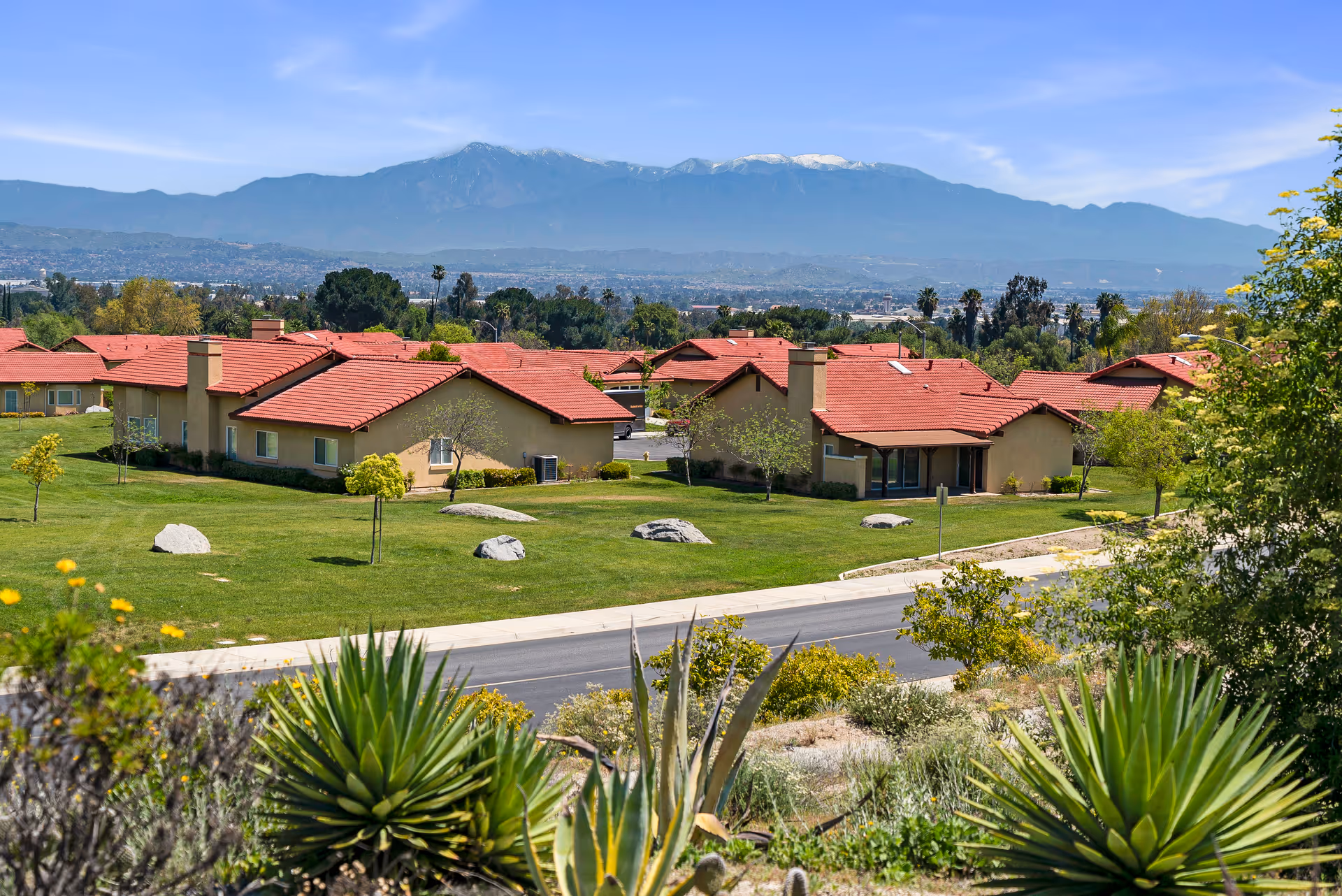 View of a senior living community with multiple single-story buildings featuring red roofs and beige walls, surrounded by green lawns, trees, and rocks. In the foreground, there are desert plants and a road. Mountains with some snow on the peaks are visible in the background under a clear blue sky.