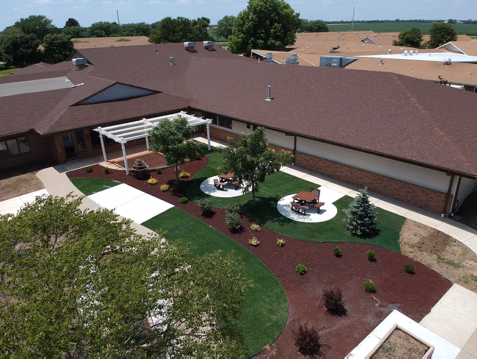 Aerial view of a landscaped courtyard at Pine Village with picnic tables, a white pergola, trees, and surrounding brick building roofs.