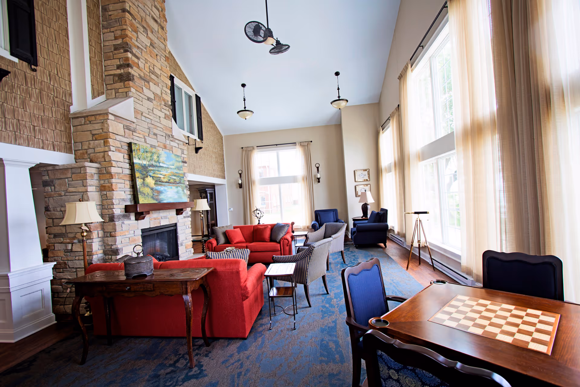 A spacious and well-lit living room with large windows covered by sheer curtains allowing natural light to fill the room. The room features a stone fireplace with a painting above it, red and blue upholstered sofas and armchairs arranged around small tables, a wooden table with a checkerboard pattern, and several lamps providing additional lighting. The walls are painted in neutral tones, and the ceiling is high with hanging light fixtures.