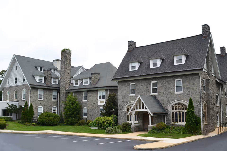 Stone-clad, multi-story senior living building with steep gabled roofs, dormer windows, an arched entrance, landscaping, and an adjacent parking area.