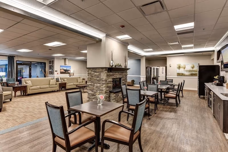 Open communal dining and lounge area with tables and chairs, a stone fireplace, sofas, and a kitchenette.
