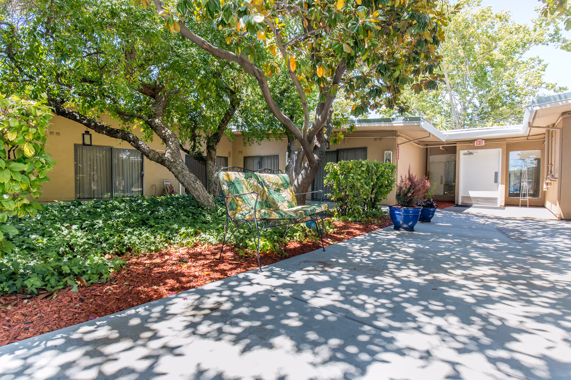 Outdoor courtyard area at Windsor Rosewood Care Center with a cushioned bench under a large tree, surrounded by green plants and shrubs. The courtyard is paved with concrete and has potted plants near the building entrance.