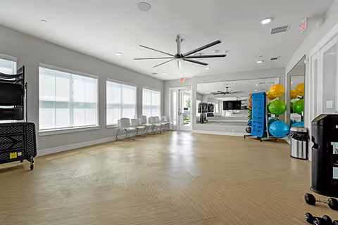 Bright multipurpose activity room with a large wall mirror, ceiling fan, row of chairs, and stacked exercise balls and equipment.