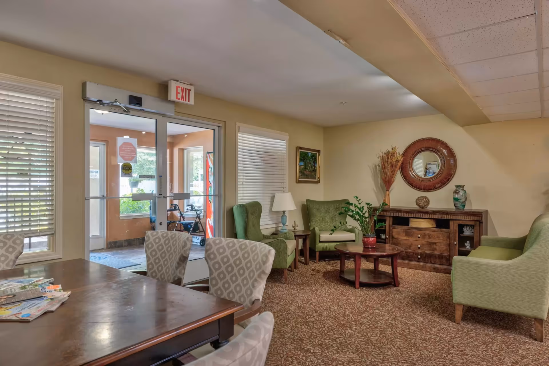 Interior view of a senior living facility common area with green upholstered armchairs, a wooden coffee table with a plant, a wooden sideboard with decorative items, a round mirror on the wall, and a table with patterned chairs in the foreground. There is a glass door with an exit sign above it and windows with blinds.
