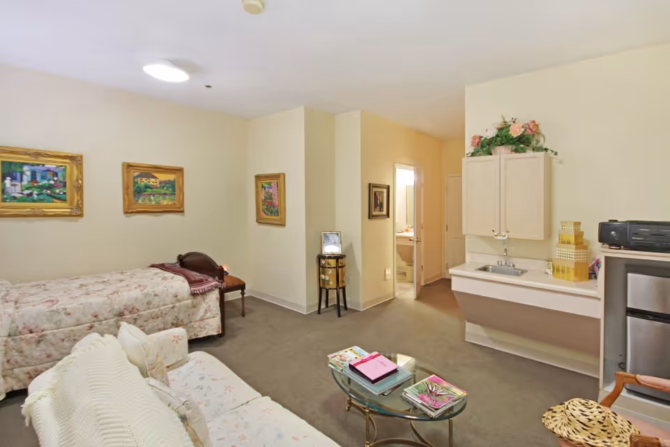 A senior living facility room with a single bed covered in a floral bedspread, a small sofa with a white knitted throw, a glass coffee table with magazines and books, a small sink with cabinets above, a mini refrigerator, and a doorway leading to a bathroom. The walls are decorated with framed paintings and there is a small table with a photo frame near the bathroom door.