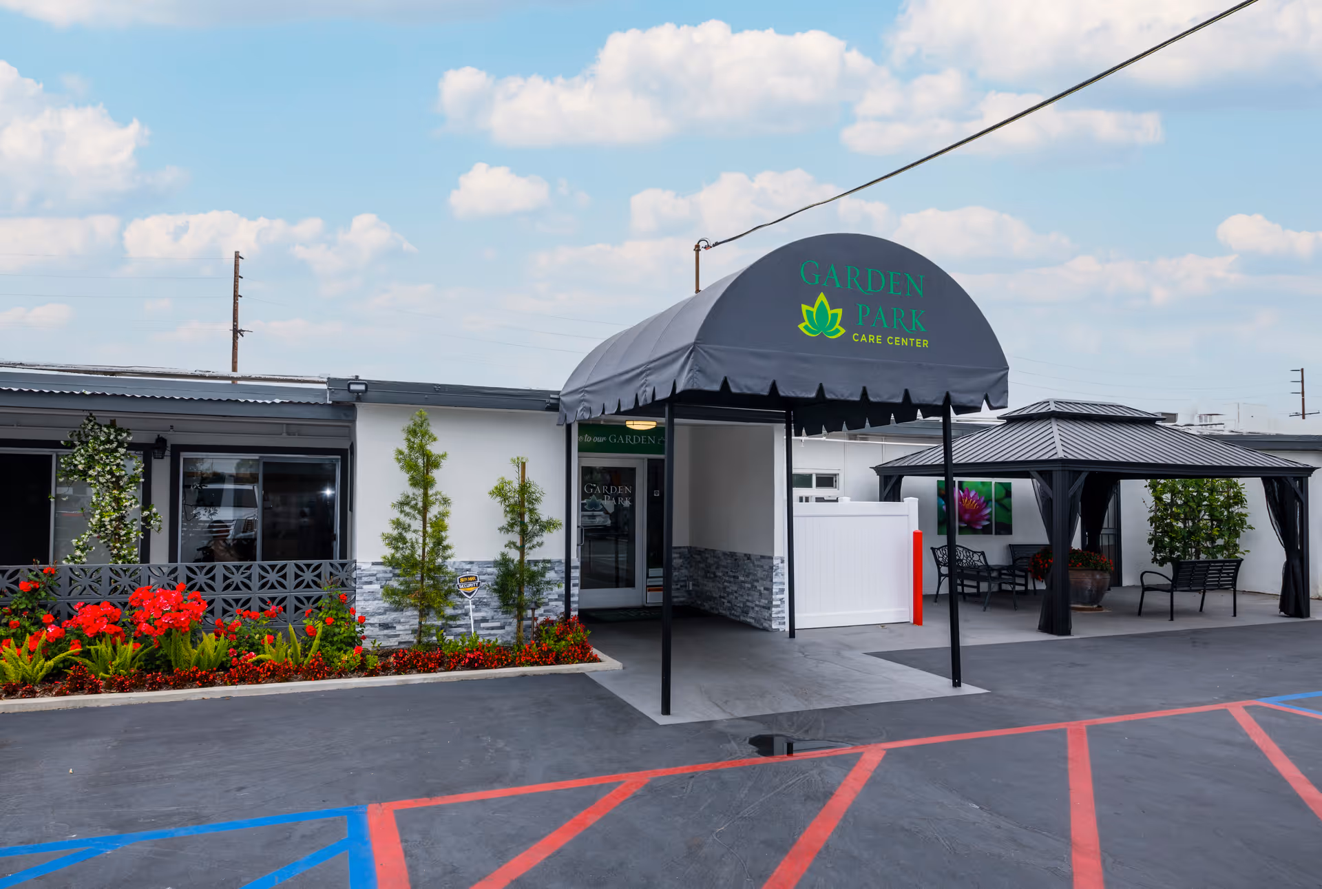 Entrance of Garden Park Care Center with a black canopy displaying the facility's name and logo. The building exterior is white with stone accents, surrounded by neatly maintained flower beds with red flowers and small trees. There is a covered seating area with black metal furniture and potted plants to the right. The parking lot in front has red and blue painted lines.