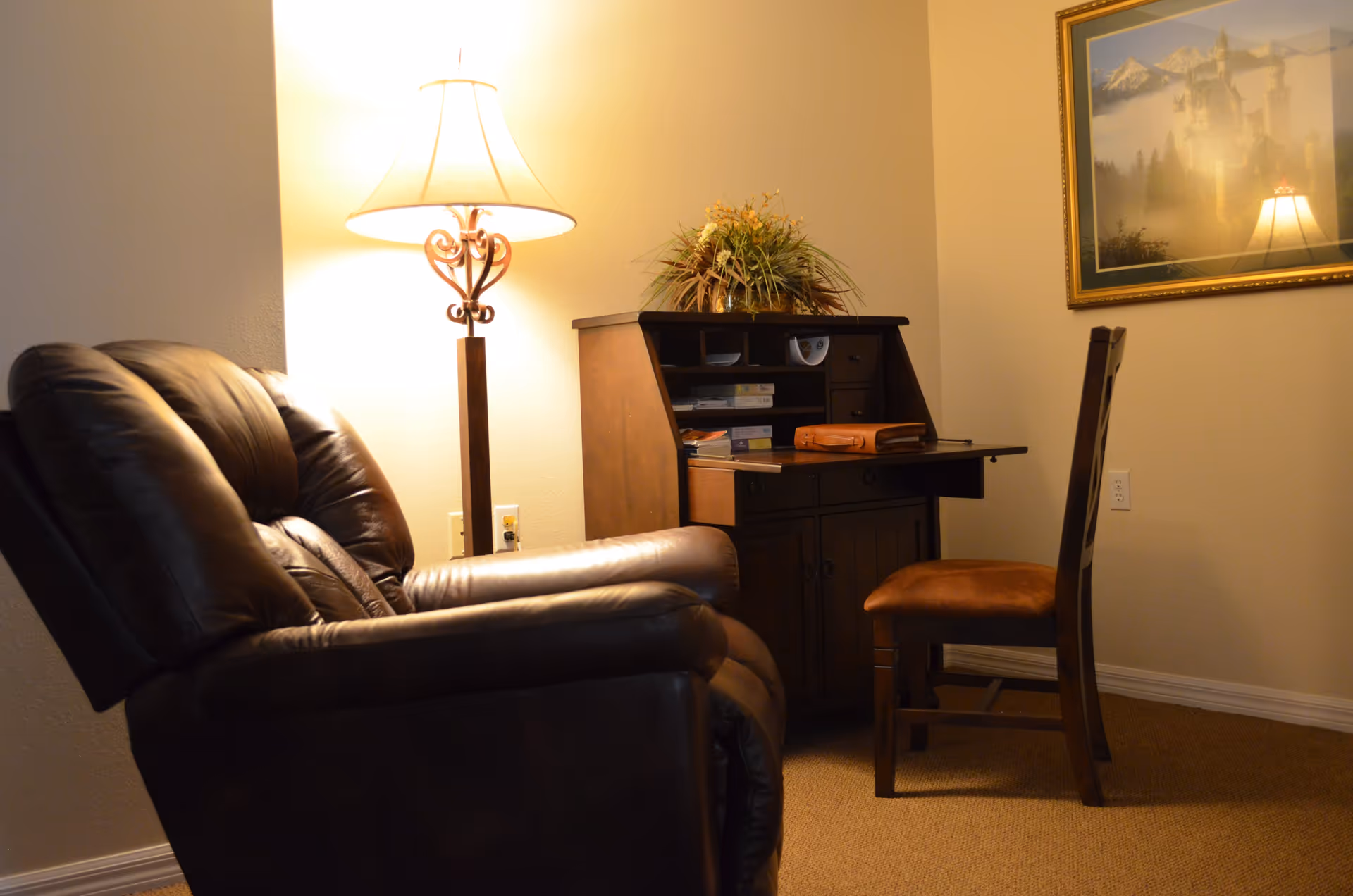 A cozy corner of a room featuring a dark brown leather recliner chair, a wooden desk with an open fold-down writing surface, a wooden chair with a cushioned seat, a table lamp with a decorative base, and a framed picture of a castle on the wall.