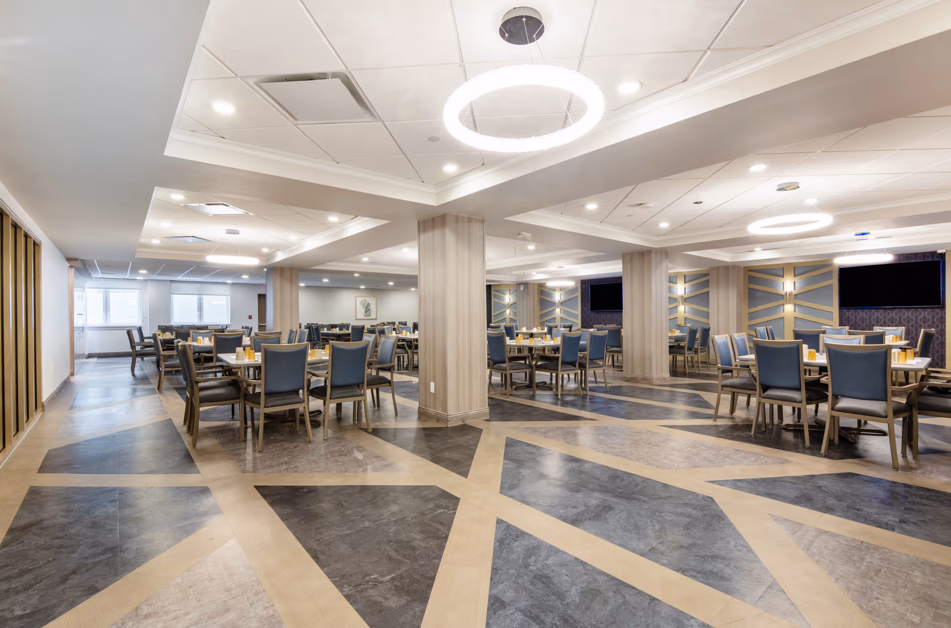 Spacious dining room in an assisted living facility with multiple tables and chairs arranged neatly. The room features modern circular ceiling lights, decorative wall panels, and a patterned floor with geometric designs.