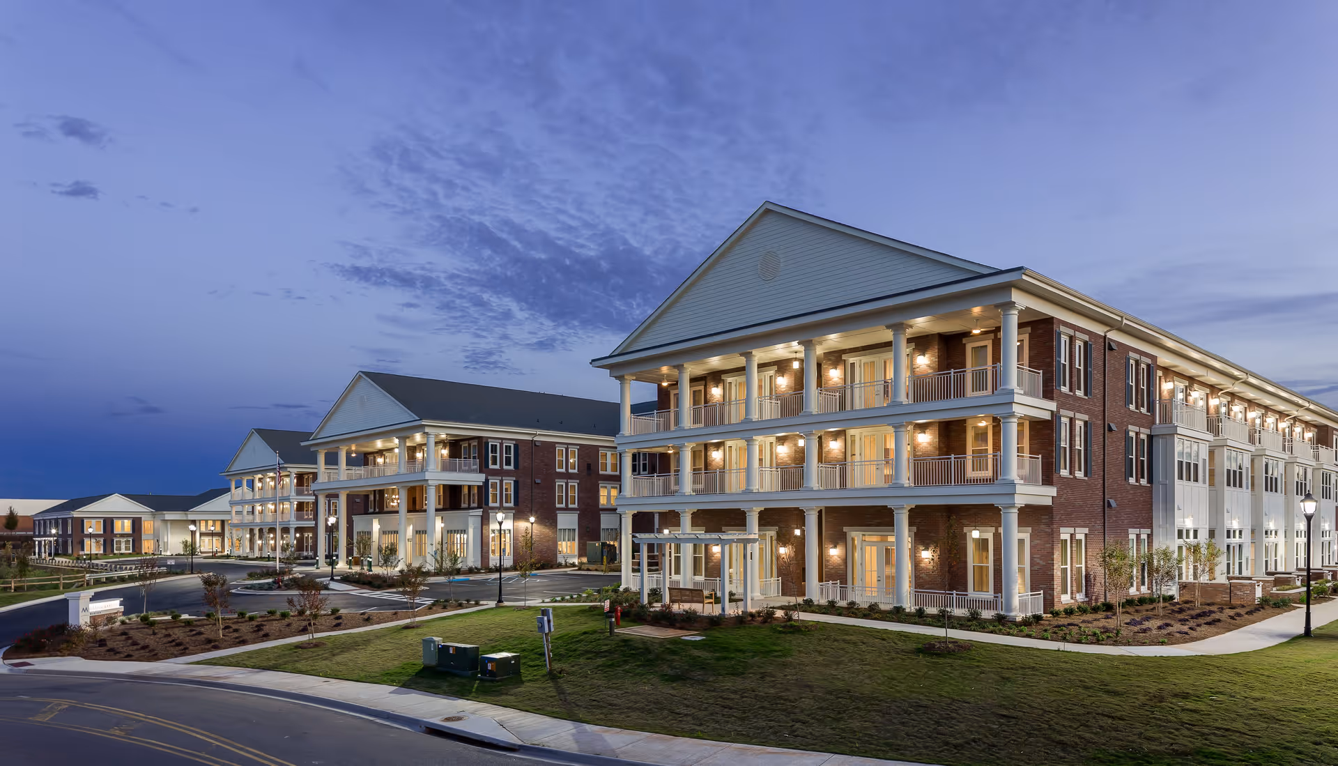 Exterior view of a large, multi-story senior living facility building at dusk with lights on in many rooms. The building features brick and white siding, multiple balconies with columns, and landscaped grounds with pathways and street lamps.