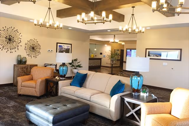 A cozy living room area in a senior living facility with a beige sofa adorned with blue pillows, two orange armchairs, a dark tufted ottoman, and side tables with blue glass lamps. The room features modern chandeliers, decorative wall art, and a mirror reflecting part of the space.