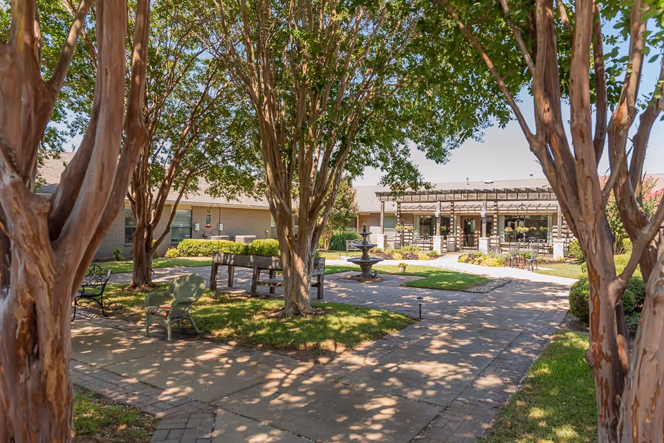 Outdoor courtyard area at Traditions Senior Living and Memory Care featuring paved walkways, several trees providing shade, benches, chairs, a central water fountain, and a building with a covered patio in the background.