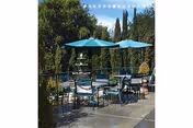 Outdoor patio area with several tables and chairs under blue umbrellas surrounded by greenery and tall trees in the background.