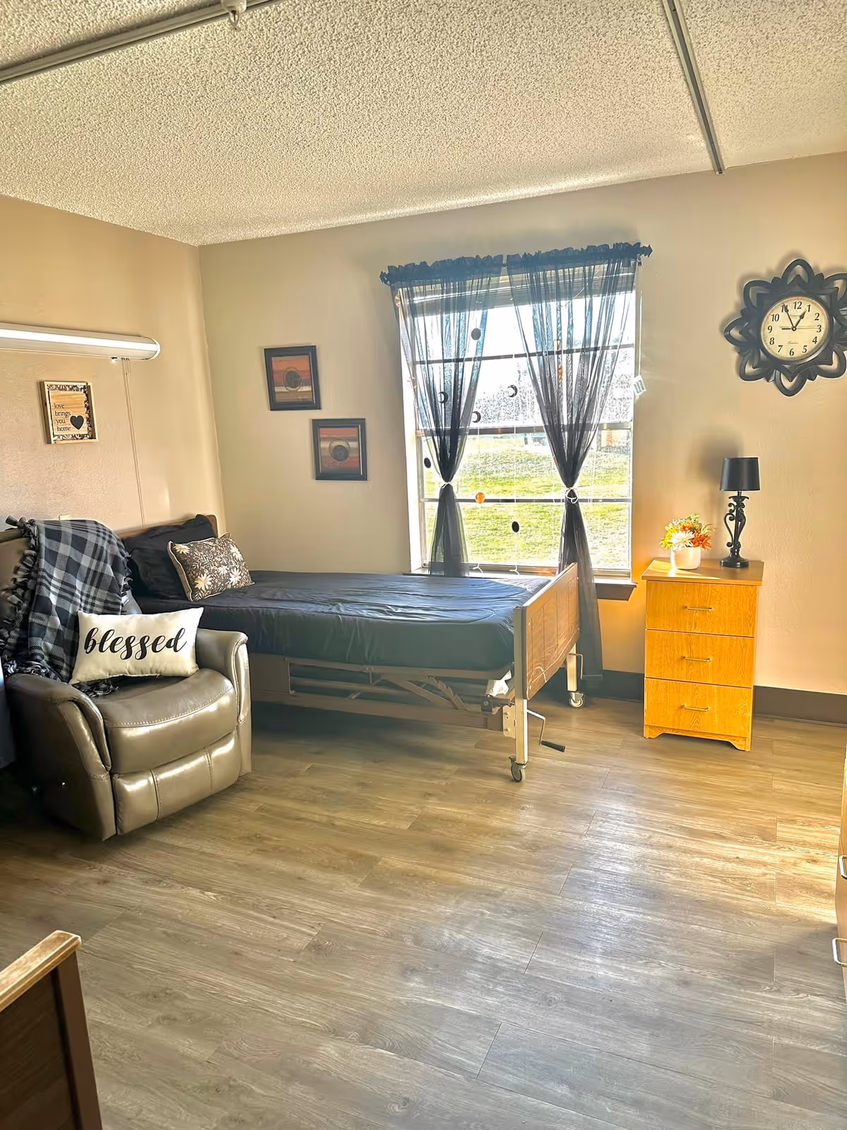 A well-lit room with a single hospital-style bed next to a window with sheer black curtains. To the left of the bed is a comfortable recliner chair with a plaid blanket and a pillow that says 'blessed'. On the right side of the bed is a wooden nightstand with a black lamp and a small flower arrangement. The walls are decorated with two framed pictures and a decorative clock. The floor is wood laminate.