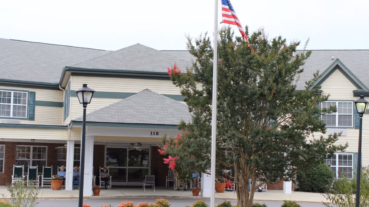 Front entrance of a senior living building with a covered porch, flagpole, large tree, and people seated outside.