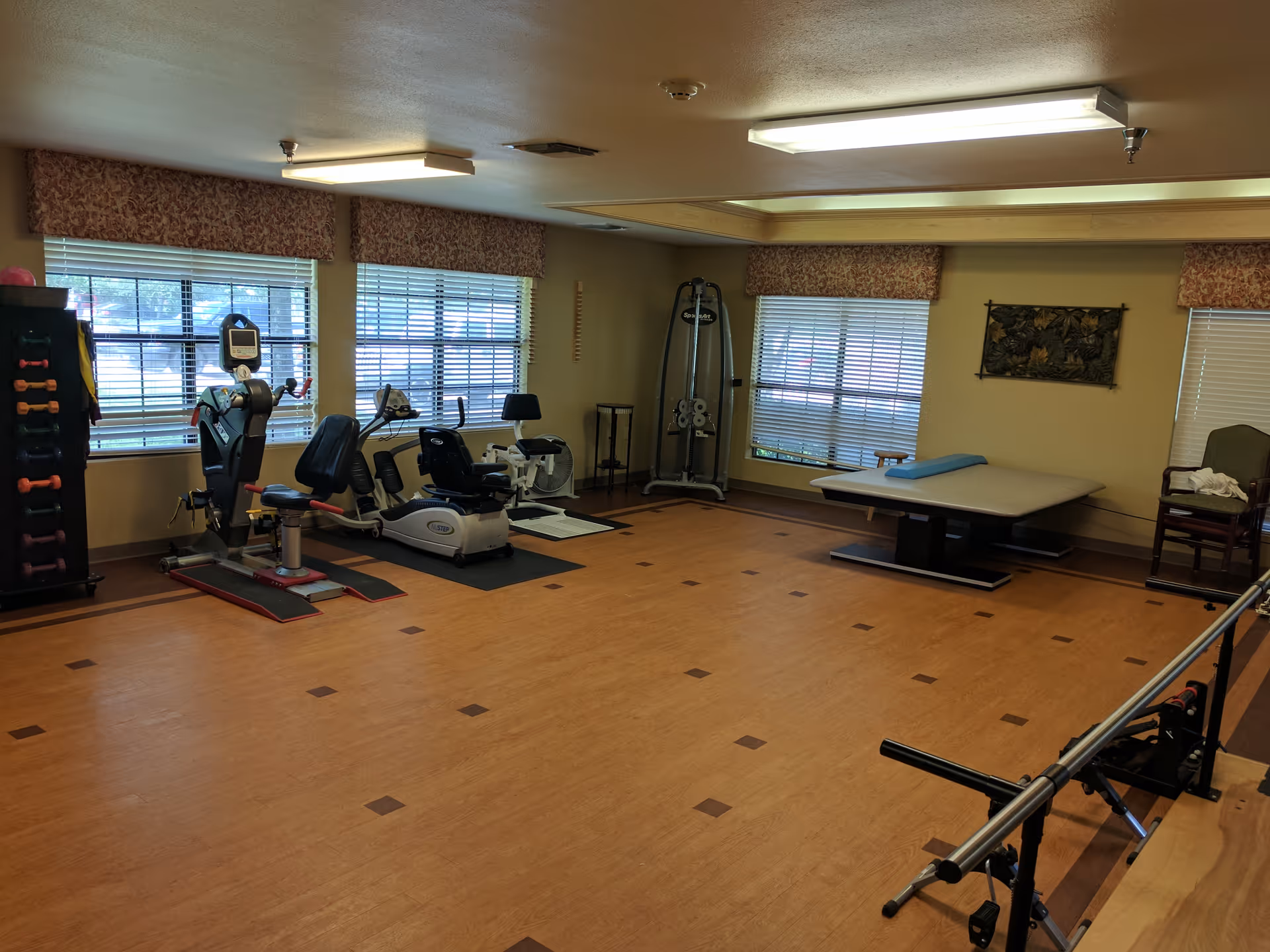A spacious exercise room with wooden flooring and large windows covered with valances. The room contains various exercise equipment including stationary bikes, a weight machine, a rack of colorful dumbbells, and a padded therapy table. There is also a chair and a small table near the therapy table. The room is well-lit with ceiling lights.