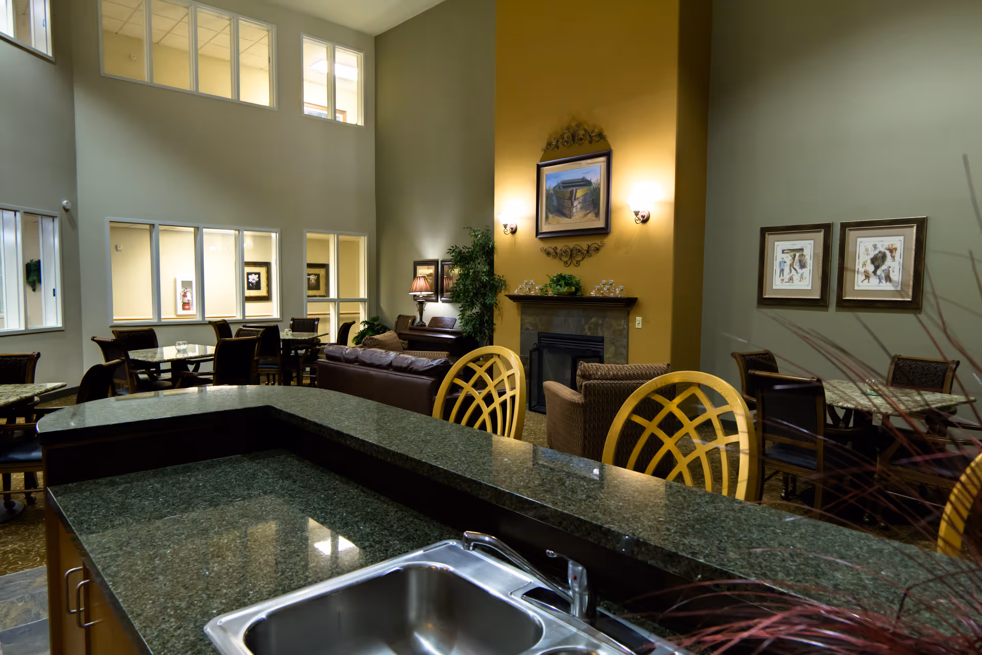 Interior view of a senior living facility common area with a granite countertop and sink in the foreground. Behind the counter are several tables and chairs, a seating area with sofas and armchairs, a fireplace with a painting above it, and wall decorations. Large windows and high ceilings provide natural light.