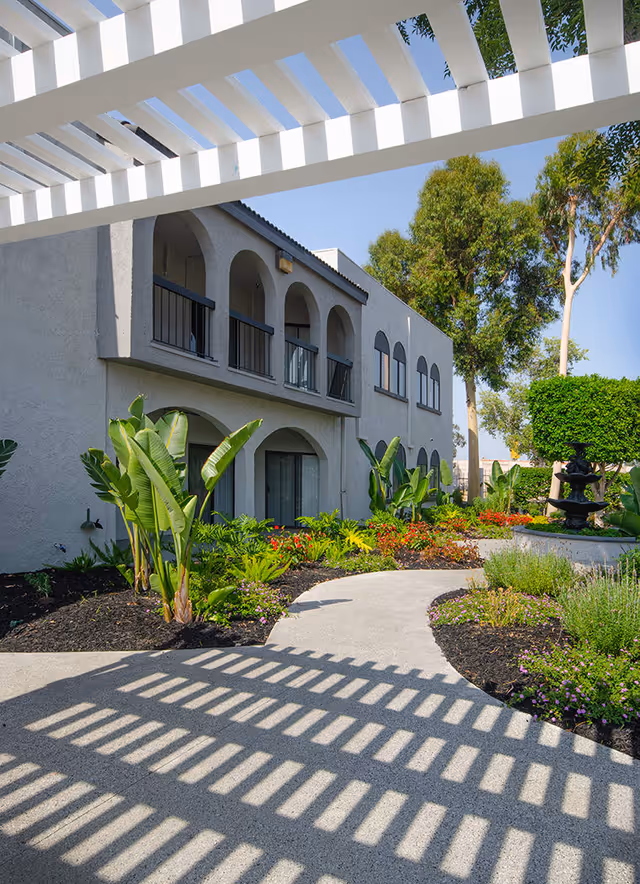 Outdoor walkway with a white pergola casting shadows on the path, surrounded by landscaped garden beds with green plants and flowers. A two-story building with arched windows and balconies is visible on the left, and trees are in the background under a clear blue sky.