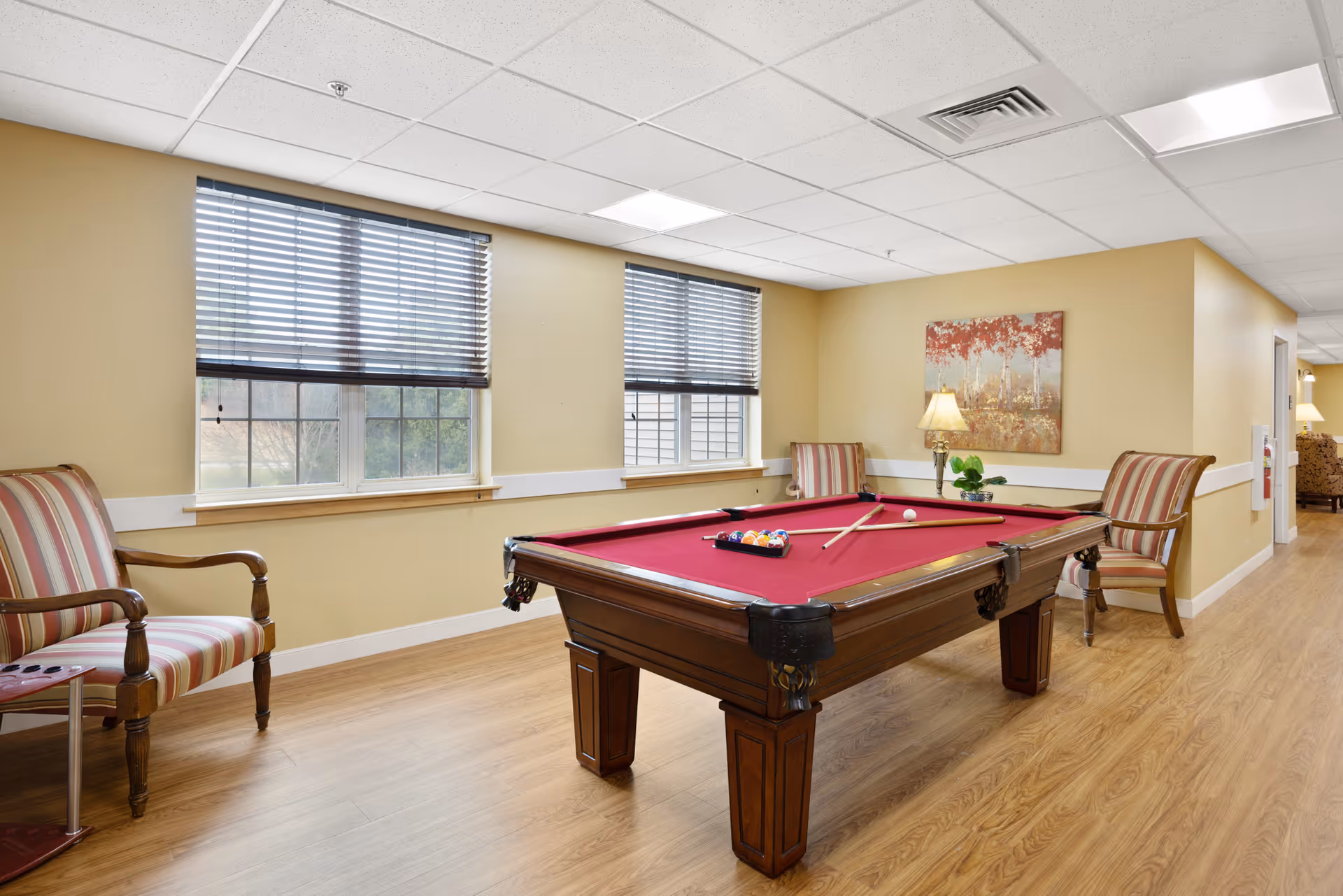 A recreational room with a red felt pool table in the center, two pool cues resting on the table, and a set of billiard balls arranged for a game. The room has light yellow walls, two windows with blinds, wooden flooring, and three striped armchairs placed around the pool table. A lamp and a painting with red and orange tones hang on the wall in the background.